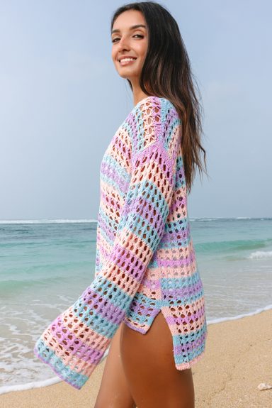 Woman smiling on a beach, wearing a colorful, loose-knit sweater by the ocean.